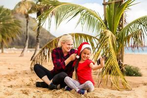 madre y hija en Papa Noel sombrero hacer un vídeo llamada por teléfono inteligente con parientes en el playa cerca palma árbol foto