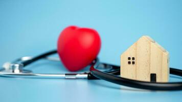 Concept of health insurance and medical welfare, small wooden house and red heart with stethoscope on blue background, health insurance and access to healthcare. photo