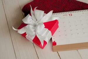 rojo regalo caja y calendario en aislado en un rojo de punto bufanda con blanco de madera antecedentes con. navidad día concepto. planificación Planificación agenda, evento, organizador Navidad día. plano poner, parte superior vista. foto