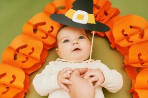 Halloween portrait of adorable baby lying on green background next to pumpkin garland, witch paper hat festive party props photo