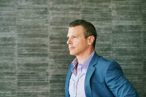 Portrait of mature 40 year old man posing bext to grey wall background, wearing purple shirt and blue jacket photo