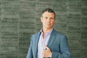 Portrait of mature 40 year old man posing bext to grey wall background, wearing purple shirt and blue jacket photo