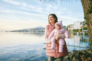 Outdoor portrait of happy young mother with adorable baby girl enjoying nice view of winter lake Geneva or Lac Leman, Montreux, Switzerland photo