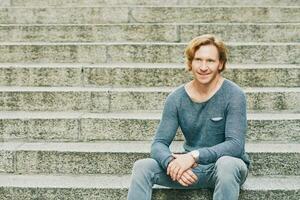 Outdoor fashion portrait of handsome red-haired man sitting on steps, wearing stylish blue pullover photo