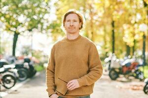 Outdoor autumn portrait of handsome young man wearing brown fuzzy fleece sweater, holding sunglasses in hands photo