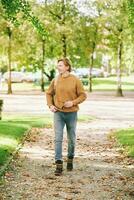 al aire libre retrato de hermoso joven hombre caminando fuera de mediante público parque, vistiendo marrón pull-over y mochila foto