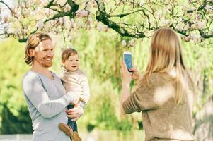 joven madre tomando teléfono imágenes de su marido y niñito hija en primavera parque foto