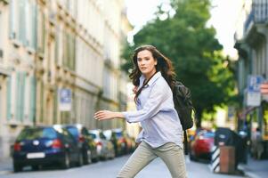 Outdoor portrait of beautiful young woman walking down the street, wearing backpack, city background photo