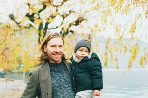 Outdoor portrait of happy young father holding toddler daughter on arms, cold autumn day in park by the lake photo