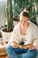 Indoor portrait of pretty young girl relaxing at home with books, female model sitting on floor, reading old vintage book, green potted plants on background photo