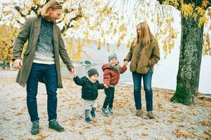 al aire libre retrato de joven contento familia de cuatro, madre y padre jugando con niños en otoño parque por el lago, frío clima foto