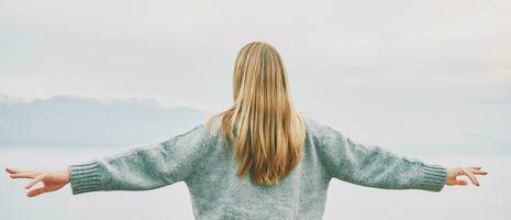 Panoramic banner background back view of young blond woman with arms wide open enjoy landscape of cloudy mountains photo