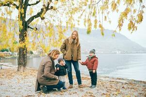 al aire libre retrato de joven contento familia de cuatro, madre y padre jugando con niños en otoño parque por el lago, frío clima foto