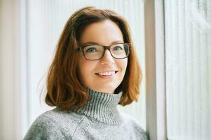 Interior portrait of beautiful 40 year old woman relaxing next to window, wearing warm grey pullover and glasses photo