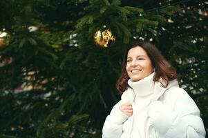 Outdoor portrait of happy woman next to green Christmas tree, wearing white pullover and jacket photo