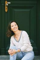 Outdoor portrait of beautiful 40 year old woman sitting on step next to green door, old money fashion style photo