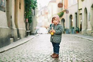 Outdoor portrait of adorable toddler girl drinking orange juice from the bottle during walk, wearing warm jacket and hat photo