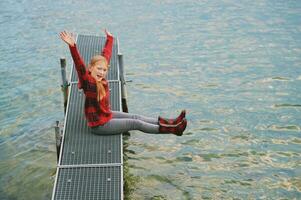 al aire libre retrato de linda pequeño niña posando siguiente a lago en muelle, vistiendo rojo suéter y lluvia botas, frío otoño o primavera clima foto