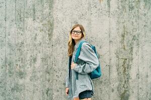 Outdoor portrait of young teenage kid girl wearing glasses and backpack, posing on grey wall background photo