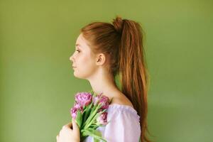 Beauty portrait of pretty young 15 - 16 year old redhaired teeenage girl wearing purple dress posing on green background, holding tulip flowers photo