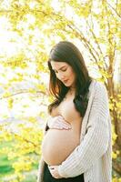 al aire libre retrato de hermosa embarazada mujer conmovedor barriga, posando siguiente a floreciente primavera flores foto