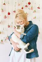 Studio portrait of pretty young teenage 15 - 16 year old girl wearing denim jacket, posing on white background with hanging flowers, holding cute cat, beauty and fashion concept photo