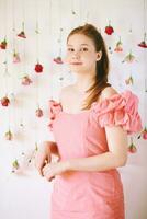 estudio retrato de bonito joven Adolescente 15 - dieciséis año antiguo Pelirrojo niña vistiendo rosado coral vestido, posando en blanco antecedentes con colgando flores, belleza y Moda concepto foto