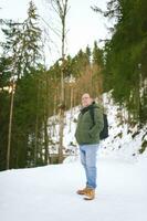 Outdoor portrait of middle age 55 - 60 year old man hiking in winter forest, wearing warm jacket and black backpack photo