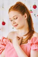 estudio retrato de bonito joven Adolescente 15 - dieciséis año antiguo Pelirrojo niña vistiendo rosado coral vestido, posando en blanco antecedentes con colgando flores, belleza y Moda concepto foto