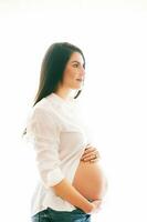 iluminar desde el fondo retrato de joven embarazada mujer, vistiendo blanco camisa y pantalones foto