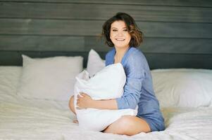 Indoor portrait of beautiful young woman relaxing in bed, holding cushion, wearing purple pullover photo