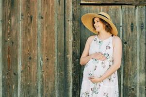 Outdoor portrait of happy young pregnant woman posing next wooden wall, wearing white dress and big hat photo