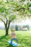 Outdoor portrait of happy young pregnant woman practicing youga in spring garden under blooming apple trees, healthy lifestyle photo