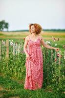 Outdoor portrait of beautiful middle age woman enjoying nice suuny evening in countryside, leaning on flower fence, wearing red dress photo