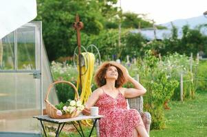 al aire libre retrato de hermosa maduro mujer descansando en verano jardín, sentado en acogedor silla, participación taza de té o café, cesta con recién cortar rosses en pie en el mesa foto