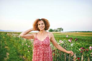 Outdoor portrait of beautiful middle age woman enjoying nice suuny evening in countryside, leaning on flower fence, wearing red dress photo