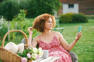 Outdoor portrait of beautiful mature woman resting in summer garden, sitting in cosy chair, having video call with her family or friend, basket with freshly cut rosses standing on the table photo