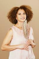 Fashion studio portrait of stylish middle age woman posing on beige background, 50 - 55 year old lady with curly hair holding pampas grass next to face photo