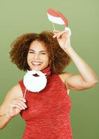 Christmas portrait of beautiful middle age woman against green background, wearing red dress, holding festive party props for photo booth