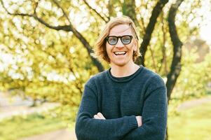 Outdoor portrait of handsome 35 - 40 year old man with red hair, posing in green sunny park, wearing blue pullover and sunglasses photo