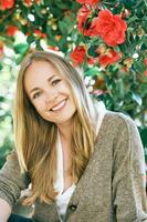 Outdoor portrait of happy young woman wearing braun jacket sitting next to red camellia flower bush photo