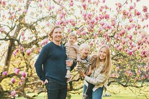 al aire libre retrato de contento joven familia jugando en primavera parque debajo floreciente magnolia árbol, encantador Pareja con dos pequeño niños teniendo divertido en soleado jardín foto