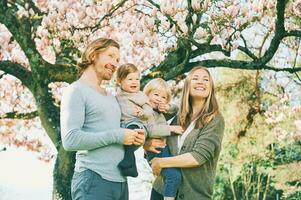 Outdoor portrait of happy young family playing in spring park under blooming magnolia tree, lovely couple with two little children having fun in sunny garden photo