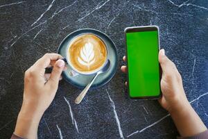 hand holding a smart phone with green screen and cup of coffee on table photo