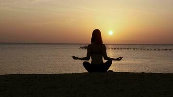 silhueta do jovem mulher relaxante e apreciar beleza pôr do sol em a oceano costa. menina fazendo ioga, meditar ao ar livre e apreciar laranja luz solar. video