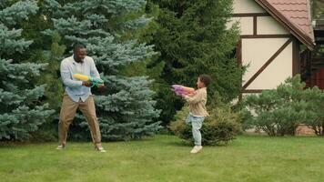 African dad and son douse each other with water guns near the house on the lawn. video