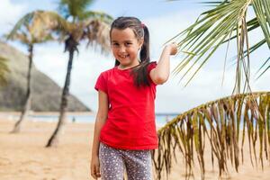 un pequeño linda bebé niña es jugando en un playa cerca un palmera. foto