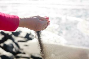 hand pours sand by the sea and rocks. symbol for time running out and time management. nails with manicure. photo