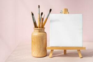 Easel with blank canvas and vase with brushes. Artistic still life. photo