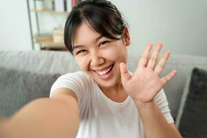 Young Asian woman using smartphone for online video conference call with friends waving hand making hello gesture photo